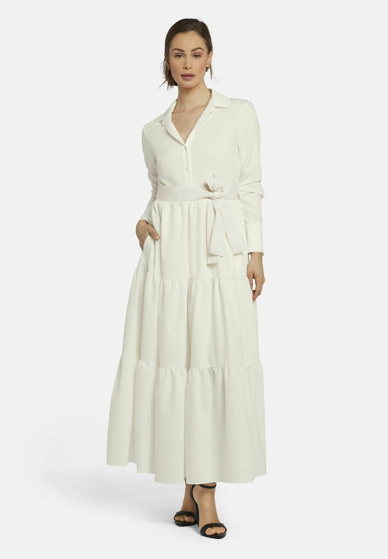 Langes weißes Kleid mit einem geknöpften Hemdkragen, Stufen am Saum, einem gebundenen Taillengürtel und seitlichen Taschen. Getragen mit schwarzen Absatzsandalen.