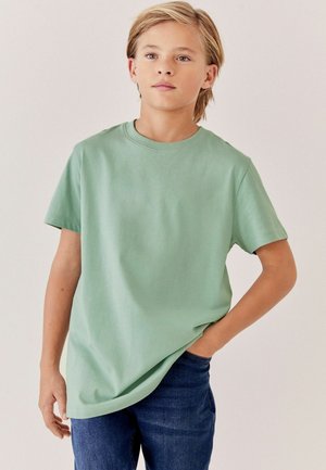 Lyshåret barn iført en lysegrøn kortærmet T-shirt og blå jeans, står med den ene hånd i lommen og kigger let til siden.