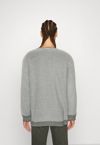 Una persona con i capelli intrecciati indossa un maglione grigio chiaro e strutturato, voltata di spalle rispetto all'osservatore su uno sfondo bianco semplice.