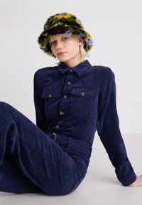 Marineblauwe corduroy jumpsuit met twee voorkruiszakken, knoopsluiting en een riem rond de taille. Multikleurige fluffy emmerhoed voegt textuur en patroon toe.