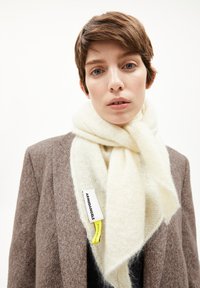 Crèmekleurige sjaal gemaakt van zacht, pluizig materiaal, met een label met zwarte en gele tekst, om de nek gedrapeerd over een bruine jas.