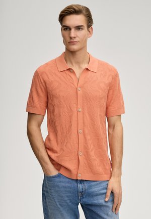 Junger Mann trägt ein korallenrotes, kurzärmeliges Hemd mit Struktur und blaue Jeans, steht mit einer Hand in der Tasche vor einem einfarbigen Hintergrund.