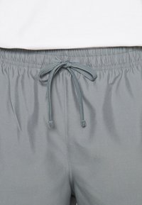 Pantalon avec taille élastique grise et cordon noué au centre, porté avec une chemise blanche rentrée au-dessus de la taille.