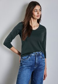 T-paita tummanvihreänä, pitkät hihat ja pyöreä pääntie, yhdistettynä vaaleansinisiin farkkuihin, joissa on klassinen leikkaus ja viisi taskua.