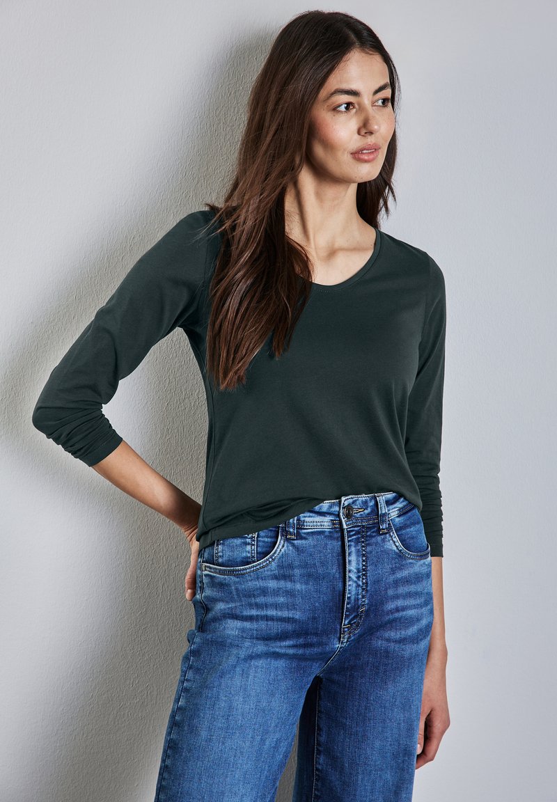 T-paita tummanvihreänä, pitkät hihat ja pyöreä pääntie, yhdistettynä vaaleansinisiin farkkuihin, joissa on klassinen leikkaus ja viisi taskua.