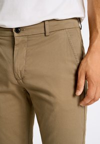 Pantalones de algodón beige con corte slim, que cuentan con bolsillos laterales, cremallera y cierre de botón negro. Textura de superficie lisa visible.