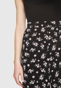 Femme portant un top sans manches côtelé noir et un pantalon noir avec un petit motif floral rose, main reposant le long du corps.