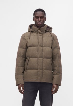 Homme portant une veste matelassée marron à carreaux avec capuche et un pantalon noir, debout devant un fond blanc uni.