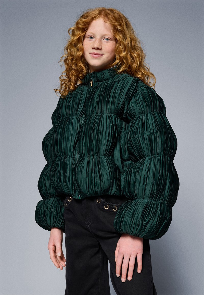 Green padded jacket with a textured finish and high collar, featuring horizontal pleats, zip closure, and elasticized cuffs. Worn with black pants.