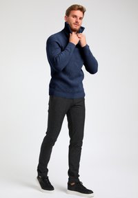 Maglione a collo alto a coste in blu navy, abbinato a pantaloni scuri a trama e sneakers nere per un look completo.