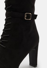 Black suede ankle boot with a high heel, featuring decorative lacing, a side strap with a gold buckle, and a smooth texture.