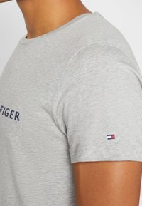 Camiseta de algodón gris con cuello redondo, que presenta un logo bordado en azul marino y un pequeño acento en rojo, blanco y azul en la manga.
