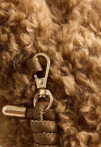 Textil marrón y esponjoso con herrajes dorados; incluye un mosquetón metálico, acentos de cuero y una correa trenzada. Detalle de superficie texturizada visible.