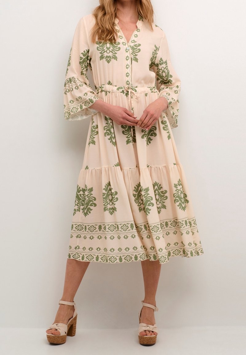Femme portant une robe midi beige avec des motifs floraux verts et des manches cloche trois-quarts, associée à des sandales compensées beiges.