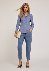 Niebiesko-biała w paski dopasowana koszula z wysmukłą kołnierzykiem, zestawiona z jasnoniebieskimi prostymi dżinsami i beżową torebką; modelka w sandałach na obcasie.