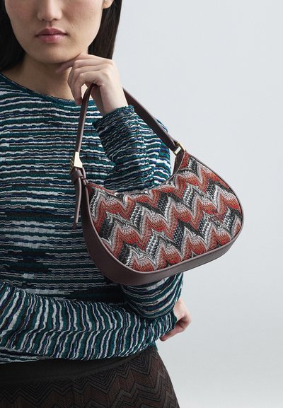 Γυναίκα κρατώντας καφέ δερμάτινη τσάντα με κόκκινο, μαύρο και άσπρο ζιγκ-ζαγκ σχέδιο, φορώντας μπλε και πράσινη ριγέ μπλούζα με μακριά μανίκια.