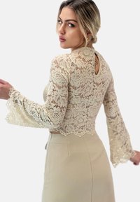 Top corto in pizzo color crema con maniche a campana, dotato di motivi floreali e un dettaglio a taglio a chiave sul retro, abbinato a una gonna beige aderente.