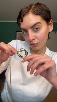 Goudkleurige hanger met een gedraaide hartvorm, bevestigd aan een fijne gouden ketting, vastgehouden tussen vingers tegen een neutrale achtergrond.