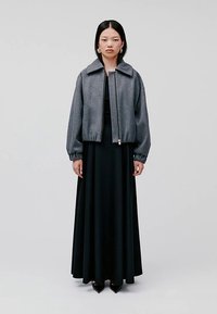 Siva pilotska jakna sa stojećim ovratnikom, patentnim zatvaračem i elastičnim manžetnama, uparena s dugom crnom haljinom i crnim šiljastim cipelama.