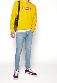 Sudadera amarilla con logo rojo de "LEVI'S", vaqueros pitillo azul claro y deportivas burdeos. Bolso con estampado de leopardo texturizado al hombro.