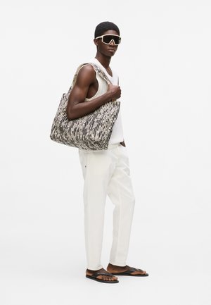 Štíhly muž v bielom nátelníku bez rukávov, bielych nohaviciach, čiernych slnečných okuliaroch a čiernych žabkách, nesúci veľkú čierno-bielu tašku s vzorom.