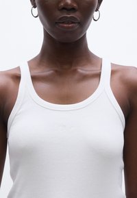 Fehér bordázott trikó vékony pántokkal és kerekített nyakkivágással. Középen kis hímzett logó található. Sima textúra.