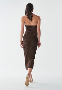 Hnedé priliehavé midi šaty s nariasenými detailmi, s viazaním za krkom a mašľou na chrbte, doplnené telovými sandálmi na podpätku.