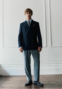 Tumus sinine topelt-rinnaga bleiser hallide volditud pükste kombinatsioonis. Riietust täiendab mustriga lips ja taskurätt, ning seda komplementeerivad mustad slip-on kingad.