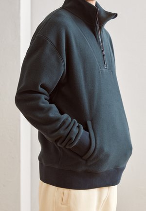 Schwarzer Fleece-Pullover mit halbem Reißverschluss, hohem Kragen, Fronttaschen, gerippten Bündchen und Saum, getragen mit hellen Hosen.