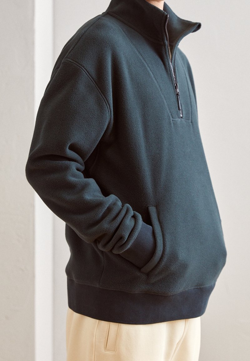 Pull en polaire noir avec demi-fermeture éclair, col montant, poches avant, poignets et ourlet côtelés, porté avec un pantalon de couleur claire.