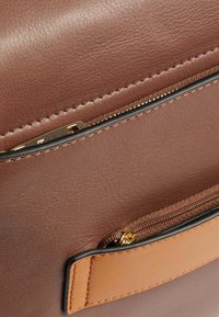 Braune Lederhandtasche mit glatter Oberfläche, ausgestattet mit einer Reißverschlusstasche mit goldenen Beschlägen und einem Akzent aus hellerem braunem Leder.