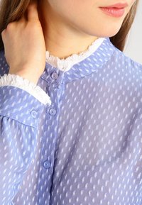 Blouse bleu clair en tissu transparent, avec un motif texturé de pois blancs, un col blanc effiloché et des boutons bleus.
