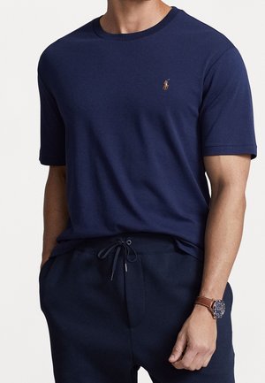 Man wearing navy blue short-sleeve crew neck shirt with small embroidered logo and matching drawstring pants, brown leather watch on wrist.