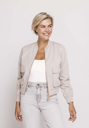 Lächelnde Frau mit kurzen blonden Haaren, die eine hellbeige Bomberjacke über einem weißen Top und hellgrauen, hoch taillierten Jeans trägt, stehend vor einem schlichten Hintergrund.