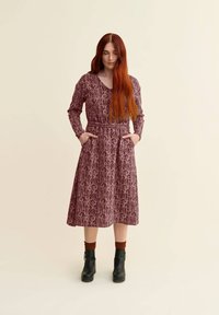 Pitkä mauve-mallinen mekko, jossa on kukkakuosi, V-aukko, pitkät hihat ja taskut. Yhdistetty mustiin nilkkasaappaisiin ja ruskeisiin sukkiin.