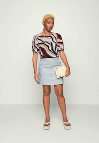 Blusa de mangas balão, multicolorida, com padrões ondulados, combinada com uma saia mini A-line azul clara. Segurando uma clutch acolchoada bege. Sandálias de plataforma verdes.