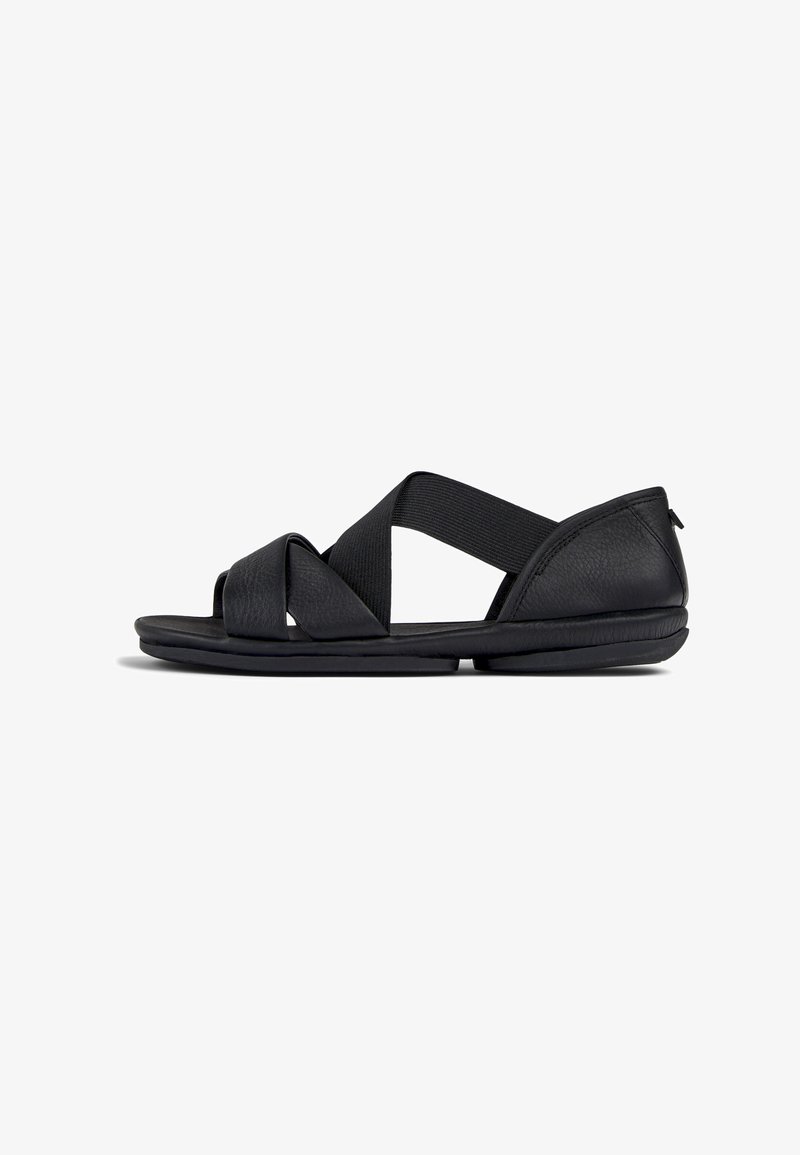 Black sandals with a textured leather upper, featuring crisscross elastic straps. The sole is flat and made of rubber for comfort.