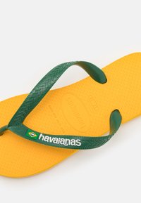 Infradito giallo in gomma con suola texturizzata, cinturini texturizzati verdi e il marchio "havaianas" in bianco. Include dettaglio con la bandiera brasiliana.