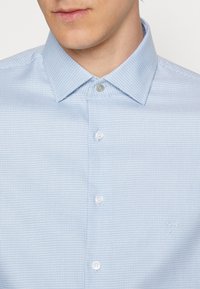 Light blue button-up shirt with a textured pattern, spread collar, and white buttons. Subtle brand logo on the lower side.