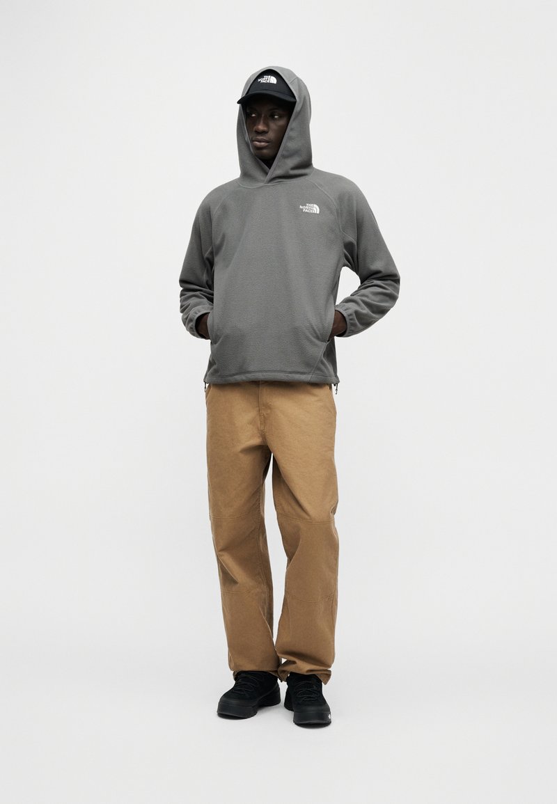 Gray fleece hoodie with a front pocket and side zippers, paired with tan pants and black shoes. The person wears a black cap.