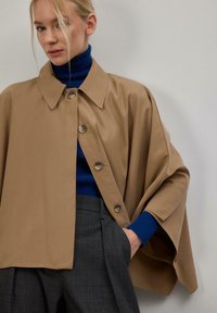 Veste cape beige avec un col à revers, cinq gros boutons et un tissu doux. Portée par-dessus un pull à col roulé bleu et un pantalon gris à carreaux.