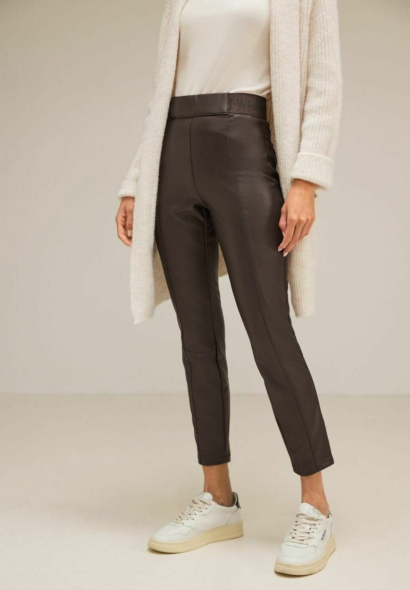 Personne portant un pantalon en cuir marron foncé taille haute, un cardigan en maille crème, un haut beige et des baskets blanches devant un fond uni.