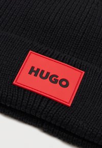 Beanie nero lavorato a maglia con texture a coste e una toppa rettangolare rossa con "HUGO" in grassetto nero.