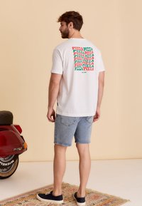 T-shirt en coton blanc avec un motif textuel coloré "PIZZA" dans le dos, associé à un short en denim clair, porté avec des chaussures navy.