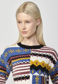 Pull en tricot avec un design en patchwork comprenant divers motifs en bleu, blanc, bourgogne, jaune et noir. Complété par un col rond noir.