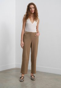 Débardeur crème clair, col V haut ; pantalon large beige avec poches avant ; chaussures noires et grises à motifs avec brides. Cadre minimaliste.