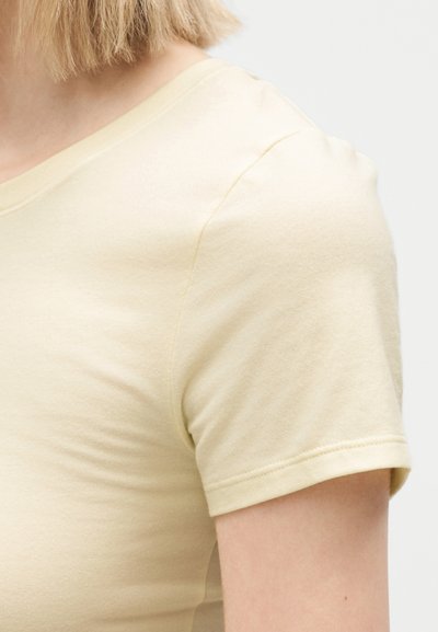 Κοντινό πλάνο ενός ατόμου που φοράει βαμβακερό μπλουζάκι με κοντά μανίκια σε ανοιχτό κίτρινο χρώμα, δείχνοντας τον ώμο και το πάνω μέρος του χεριού μπροστά σε μονόχρωμο φόντο.