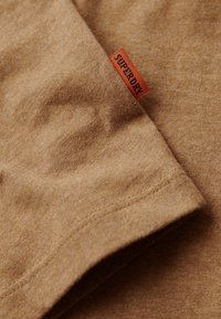T-shirt di cotone marrone con una consistenza morbida e un'etichetta arancione "SUPERDRY" cucita di lato. Presenta un design semplice e una finitura liscia.