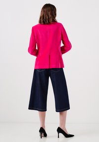 Chaqueta de terciopelo fucsia con mangas largas y una abertura trasera, combinada con pantalones de mezclilla oscuros de pierna ancha y tacones negros de punta.