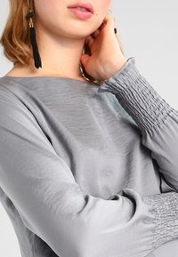 Tessuto grigio lisciato con scollo ampio e dettagli a manica arricciata. Il modello mostra una vista laterale, evidenziando la texture e il drappeggio delle maniche.